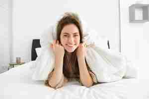 Free photo cute girl with messy hair lying in bed covered in white sheets duvet smiling and laughing coquettish