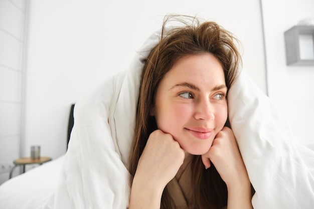 Cute girl with messy hair lying in bed covered in white sheets duvet smiling and laughing coquettish