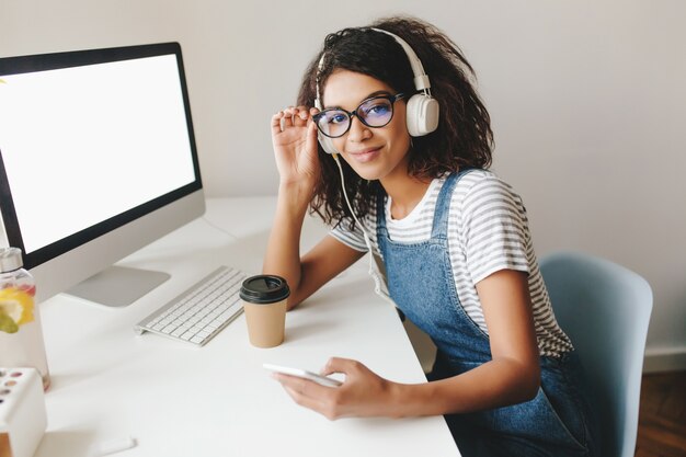 Cute girl with tired smile posing at workplace near computer with white screen