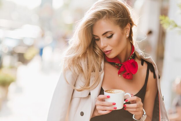 Cute girl with trendy make-up relaxing in sunny day and drinking latte with eyes closed. Outdoor portrait of gorgeous tanned woman with blonde hair posing in coat with cup of coffee.