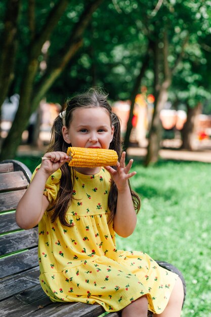 A cute girl in a yellow dress is sitting on a bench and eating a yellow corn cob in an amusement par...