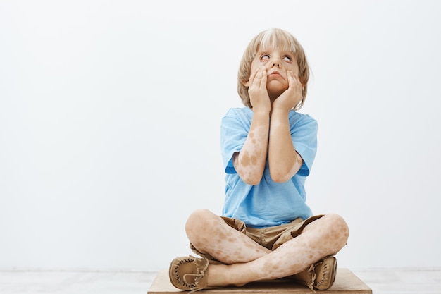 Free photo cute good-looking child with two-colored skin, sitting with crossed feet, rolling eyelids and pulling eyes with hands, feeling bored while being punished