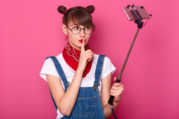 cute good looking young woman with thoughtful expression, cons mouth with fore finger, keeps secret, being photographed by hersrelf, isolated on pink. Teens concept