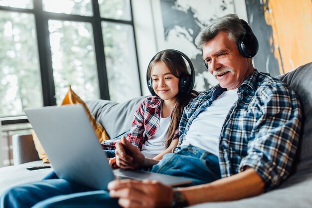 Милый внук и ее дедушка слушают музыку на цифровом планшете, сидя на диване.