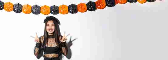 Бесплатное фото Милая счастливая азиатка, наслаждающаяся вечеринкой в честь хэллоуина, показывающая мирный жест и улыбающаяся в ведьминской стоимости