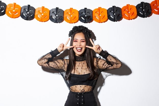 Cute happy asian woman enjoying halloween party, showing peace gesture and smiling, wearing witch costume, standing against pumpkin banners decoration. Free Photo