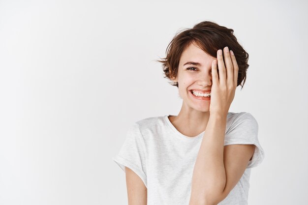 Cute and happy caucasian woman in t-shirt, cover half face and smiling , standing against white wall