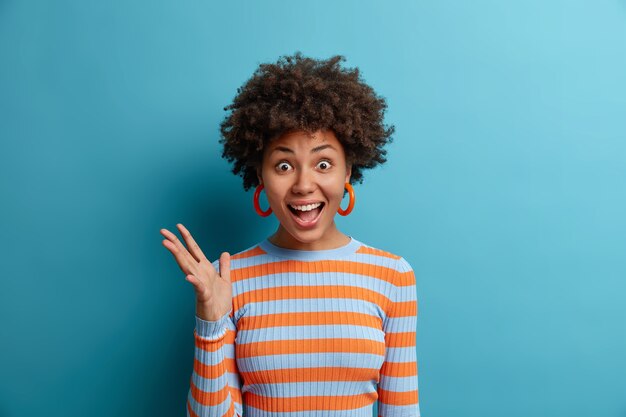 Cute happy curly woman looks full of disbelief, raises hand and exclaims with joy, gets excellent news or pleasant gift dressed casually isolated on blue wall. Positive human reaction, emotions