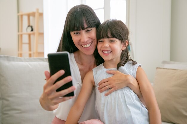 Cute happy girl and her mom using phone for video call while sitting on sofa at home together