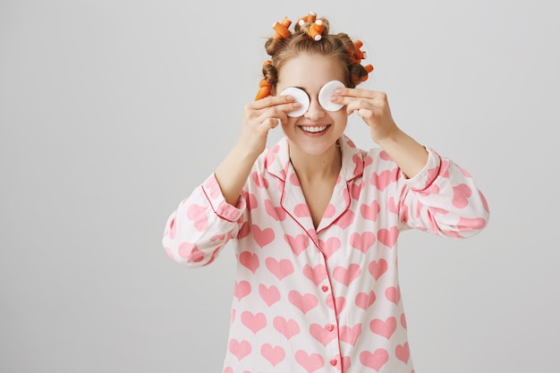 Free photo cute happy girl in nightwear and hair curlers wipe-off makeup with cotton pads