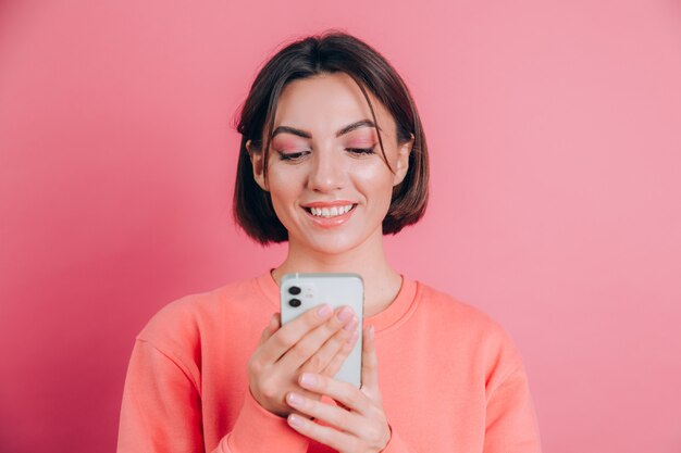 Cute happy young beautiful woman posing isolated over pink wall background using mobile phone