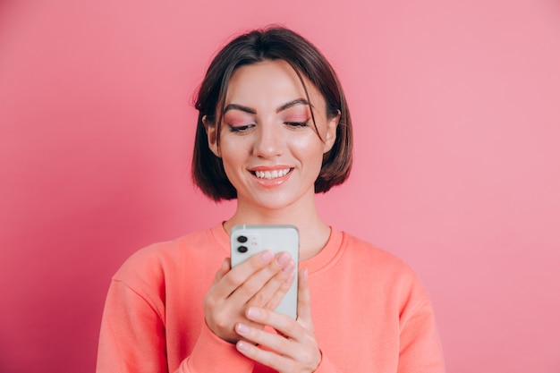 Милая счастливая молодая красивая женщина позирует изолированной на розовом фоне стены с помощью мобильного телефона