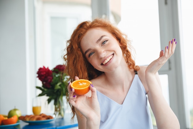 Cute happy young redhead lady near flowers holding orange