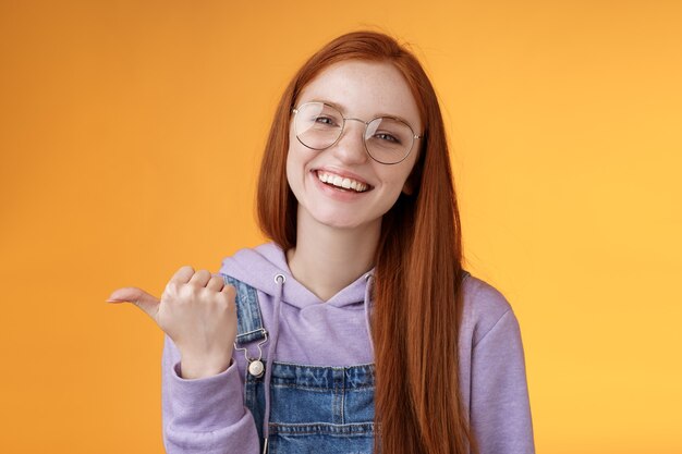 Cute helpful friendly-looking joyful european redhead woman show thumb left smiling delighted laughing pointing where find awesome store telling about interesting new promo offer, orange background