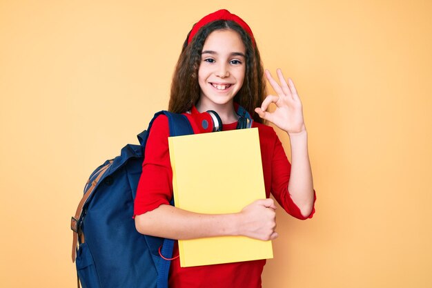 Cute hispanic child girl wearing student backpack and headphones holding book doing ok sign with fingers smiling friendly gesturing excellent symbol