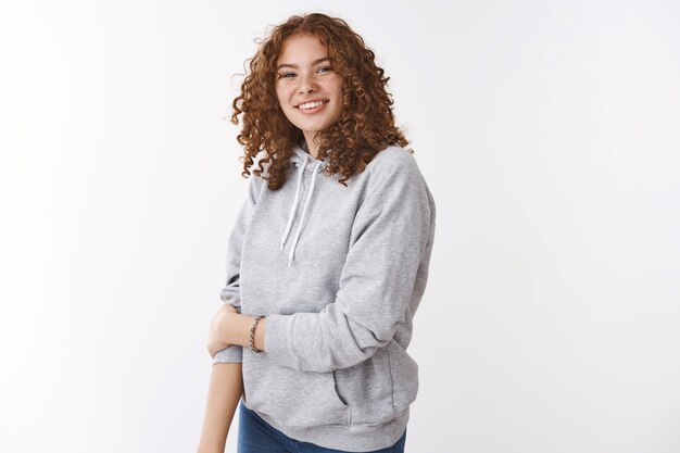 Cute insecure young redhead teenage girl with freckles curly hair wearing grey hoodie smiling touch arm unconfident blushing grinning friendly talking with new coworkers, standing white background