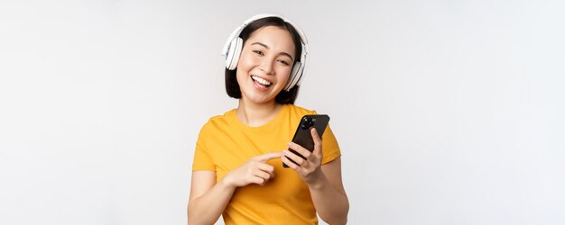 携帯電話を見て、白い背景に立っているスマートフォンの音楽アプリを使用して笑っているヘッドフォンでかわいい日本人の女の子