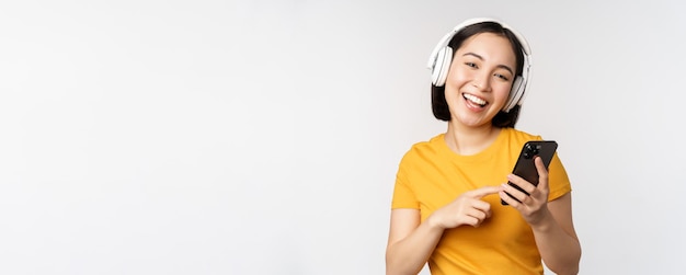 Cute japanese girl in headphones looking at mobile phone and smiling using music app on smartphone standing against white background