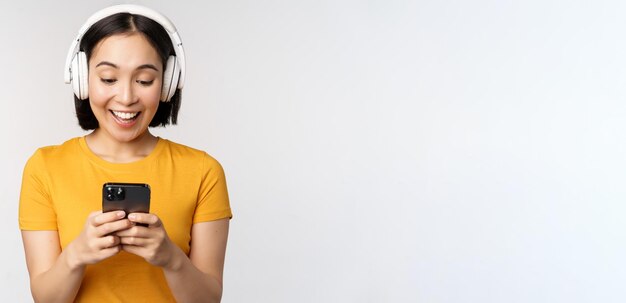 Cute japanese girl in headphones looking at mobile phone and smiling using music app on smartphone standing against white background