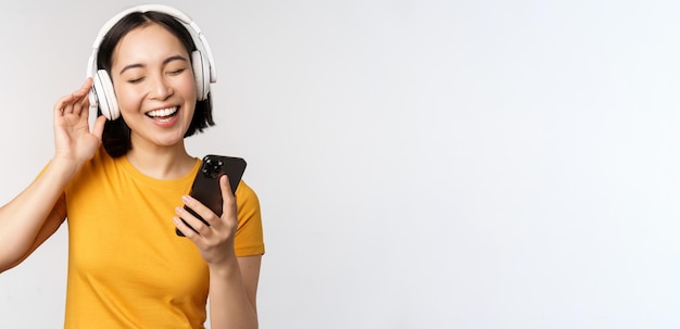 헤드폰을 끼고 휴대폰을 보고 흰색 배경에 서서 스마트폰의 음악 앱을 사용하여 웃고 있는 귀여운 일본 소녀