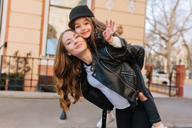 Cute joyful girl in black hat waving hand, riding on mother's back during walk around city. Outdoor portrait of lovely woman in trendy jacket carrying daughter and posing in front of building.