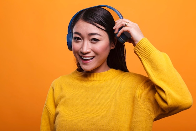 かわいいうれしそうなポジティブなアジア人は、オレンジ色の背景にいる間、仲間と話すために音楽を聞くのをやめます。彼女は彼女の友人に会うときにヘッドフォンを脱いで美しい笑顔の心のこもった女性。