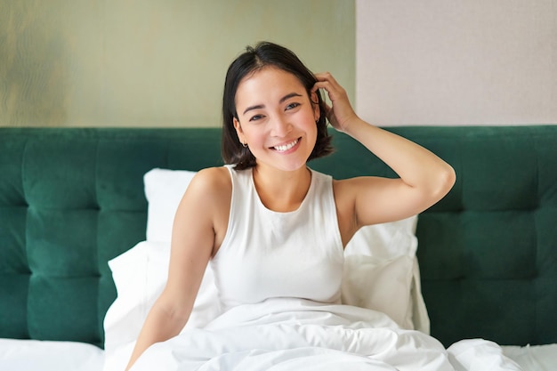 무료 사진 흰색 탱크 탑을 입은 귀여운 한국 소녀는 아침 스트레칭 손에 침대에 누워 그녀의 침실에서 깨어납니다.