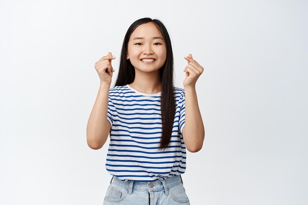 Cute korean girl shows finger hearts gesture and smiling optimistic and positive face expression happy attitude standing over white background