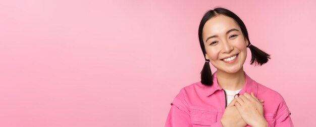 Cute korean girl with silly hairbuns looking happy and grateful thank you pose holding hands on heart flattered posing against pink background