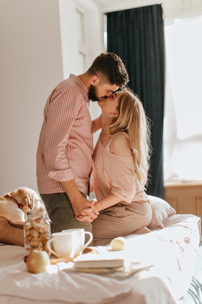 귀여운 래브라도가 침대에 누워 있는 동안 그의 주인은 아름다운 아침을 즐기고 아침 식사 중에 침실에서 로맨틱한 커플의 스냅샷에 키스합니다