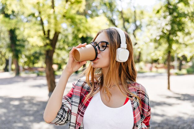 路上で喜んでコーヒーを飲むかわいい女性。木の前に立っている白いヘッドフォンで魅力的な女性モデル。