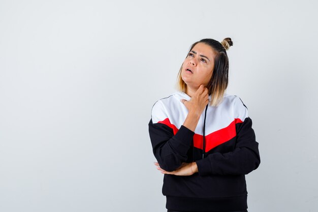 Cute lady looking up, touching neck in hoodie and looking pensive , front view.