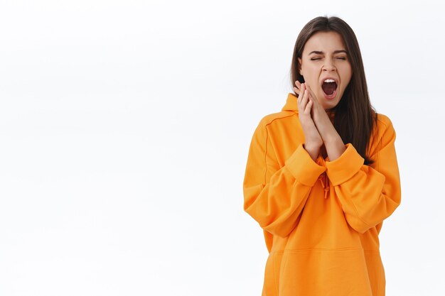 Cute lazy brunette girl in orange hoodie close eyes and yawning with opened mouth, feeling sleepy, wake up morning want coffee, bored of uninteresting conversation, white wall