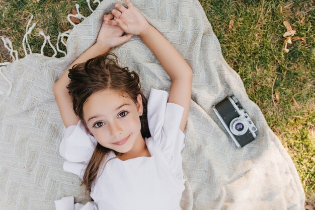 Cute lightly tanned girl with shiny beautiful eyes posing on blanket with camera during summer weekend. Overhead portrait of brown-haired female kid lying on the grass and dreaming.