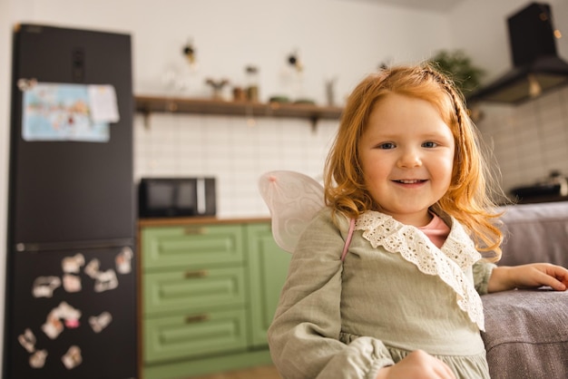 Free photo cute lightskinned little redhead girl in dress looks at camera while standing in kitchen concept of children's lifestyle