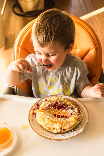 Симпатичный маленький ребенок ест блин на тарелке над высоким стулом