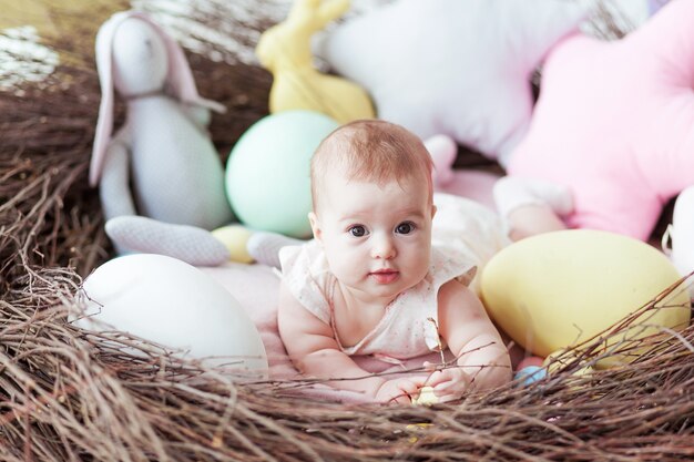 Cute little baby with colorful Easter eggs and toy rabbit lying in nest.