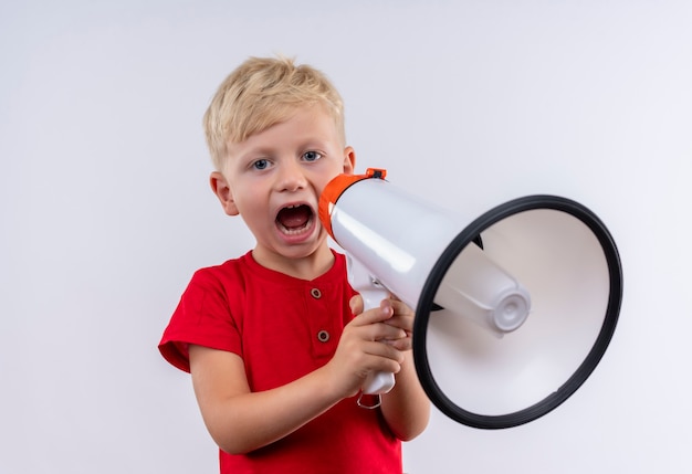 A cute little blonde boy wearing red t-shirt speaking through megaphone while looking on a white wall