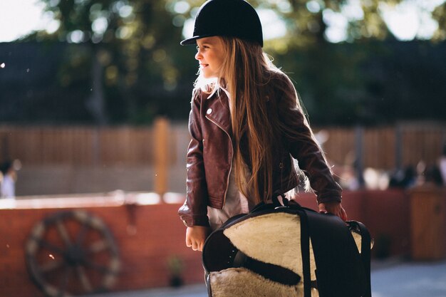 Cute little blonde girl with saddle in a stable