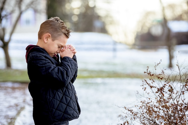 Милый маленький мальчик молится с закрытыми глазами посреди зимнего парка
