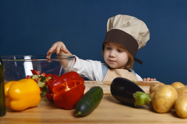 милый маленький мальчик в шляпе шеф-повара стоит за кухонным столом