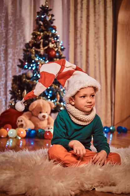 家のクリスマスツリーの近くの毛皮のカーペットの上に座っている装飾的な帽子とスカーフを身に着けているかわいい男の子。