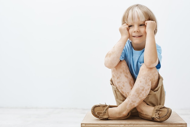 Cute little boy with blond hair and spots on skin, sitting with crossed feet, holding hands near face and smiling with joyful carefree expression, looking aside