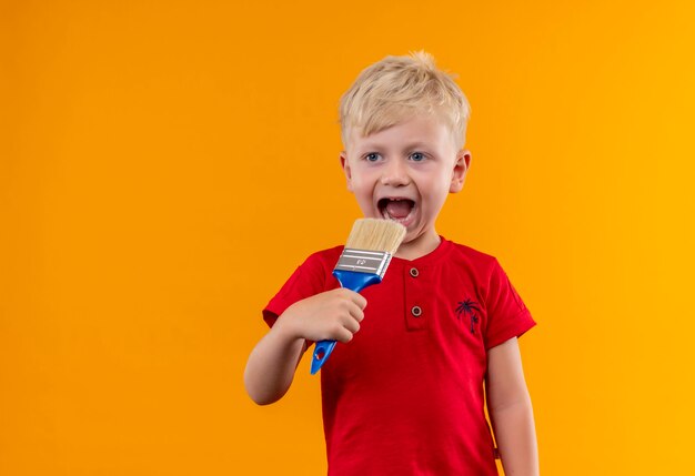 A cute little boy with blonde hair and blue eyes wearing red t-shirt holding blue paint brush close to mouth looking side on a yellow wall