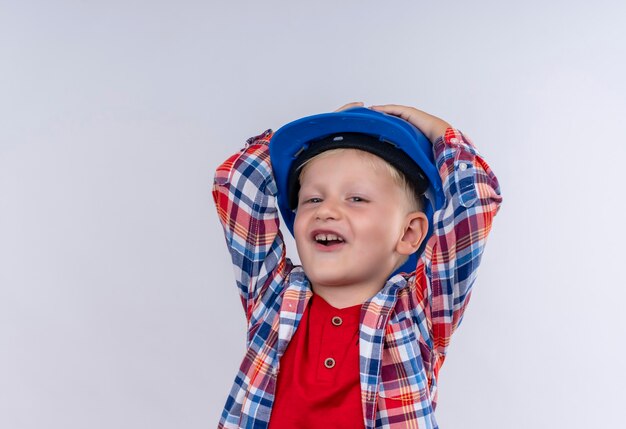 흰 벽에 그의 머리에 손을 잡고 파란 헬멧에 체크 셔츠를 입고 금발 머리를 가진 귀여운 어린 소년