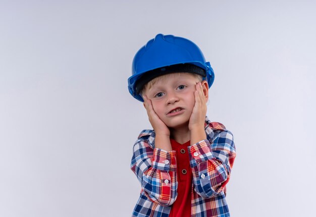 Симпатичный маленький мальчик со светлыми волосами в клетчатой рубашке в синем шлеме, держа руки на лице на белой стене