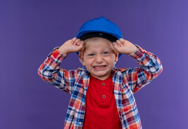 A cute little boy with blonde hair wearing checked shirt in blue helmet raising clenched fists looking on a purple wall