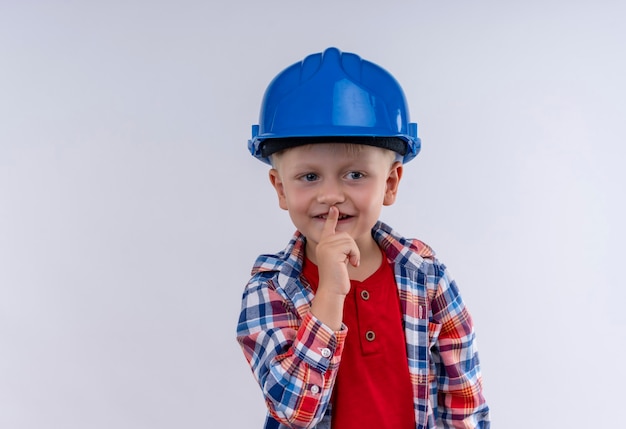 A cute little boy with blonde hair wearing checked shirt in blue helmet showing shh gesture on a white wall