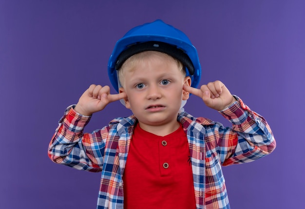 紫色の壁を見ながら耳に指を保ちながらヘルメットにチェックのシャツを着ているブロンドの髪のかわいい男の子