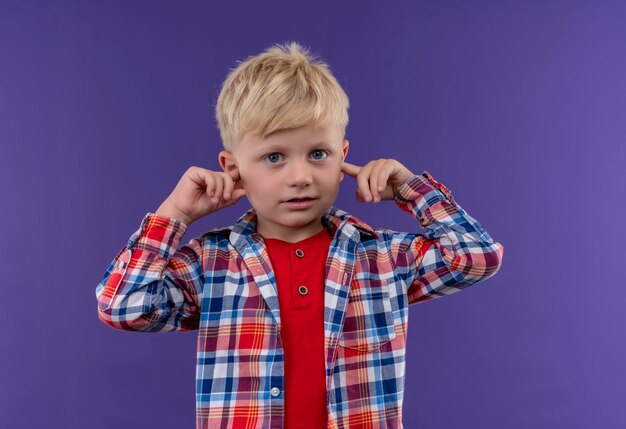 A cute little boy with blonde hair wearing checked shirt keeping hand on ears on a purple wall